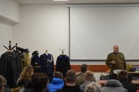 sala konferencyjna, siedzi grupa osób przed którą stoi mężczyzna w mundurze policyjnych z lat 20-tych, po prawej stornie na manekinach rozwieszone części mundurów z tego samego okresu