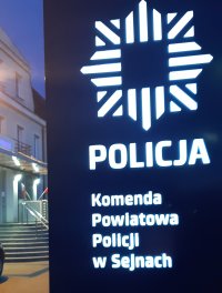 na pierwszym planie tablica z napisem Komenda Powiatowa Policji w Sejnach, w tle wejście do budynku komendy